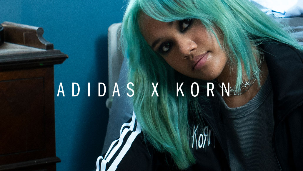 Eine Frau mit türkisen Haaren blickt in die Kamera und trägt eine schwarze adidas x Korn Jacke (Foto)