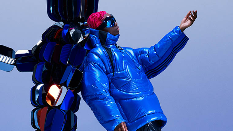 Eine Person mit roten Haaren und einer großen Sonnenbrille trägt eine adidas x Moncler Daunenjacke und lehnt sich an eine Figur aus blauen Würfeln (Foto)