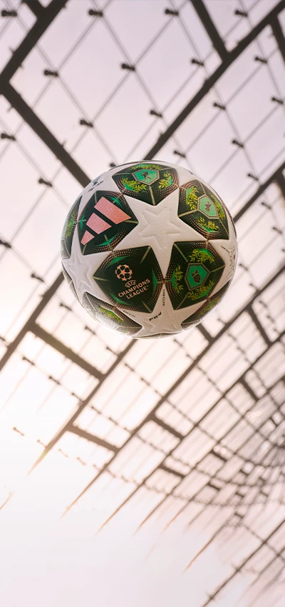 This image shows an interior view of what looks like a soccer stadium with perforated metal mesh walls and ceiling. In the foreground, there is a patterned ball marked with the stars of the UEFA Champions League.
