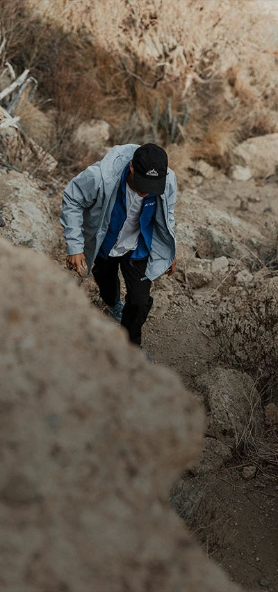 The image shows a person standing in what looks like an outdoor, possibly natural setting. The individual is bending down with one hand on their hip and the other extended outwards, as if they are inspecting something or perhaps posing for the photograph. They are wearing casual attire consisting of pants and a long-sleeve shirt, along with some kind of cap or head covering.