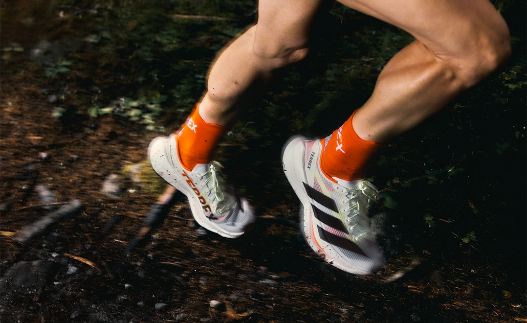 Nahaufnahme von Beinen beim Trailrunning im Wald, getragen werden adidas TERREX Laufschuhe mit drei Streifen und orangefarbenen Socken. (Foto)
