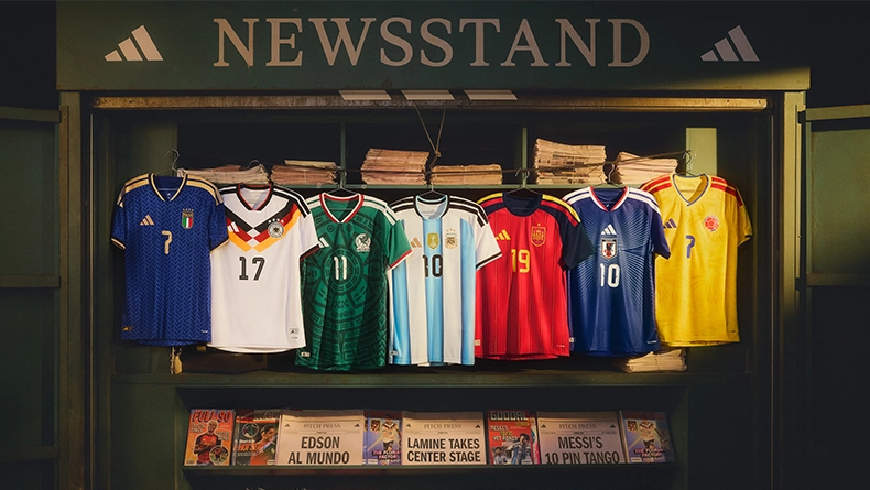 Fußballtrikots verschiedener Nationalteams hängen in einem beleuchteten Kiosk mit der Aufschrift „Newsstand“, darunter liegen Zeitungen und Magazine. (Foto)