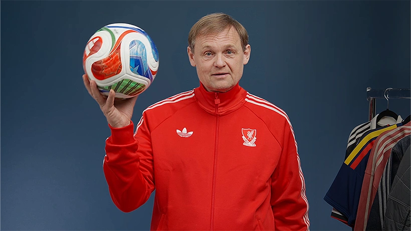 adidas CEO Bjørn Gulden wearing a red adidas tracksuit holding a football, with jerseys hanging on a rack beside him. (Photo)